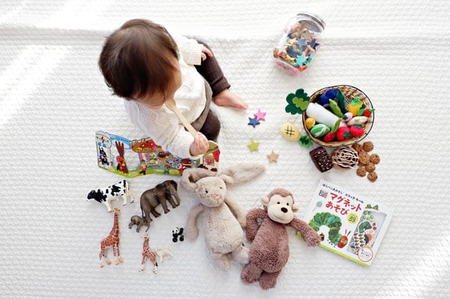 Cómo organizar todos los artículos para bebé en casa, a baby sitting on a playmat surrounded by stuffed animals, toys, and cardboard baby books.